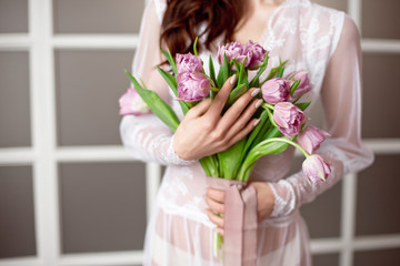 Beautiful young woman in the white dress with flowers tulips in hands on bed at home.