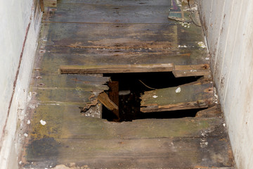 A hole in the floor of an old building. The floor is in a narrow hallway. A dark basement can be...
