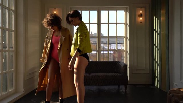 Two women in bathing suits and outerwear pose for the camera in full growth. Fashion shooting in the Studio with large Windows and parquet