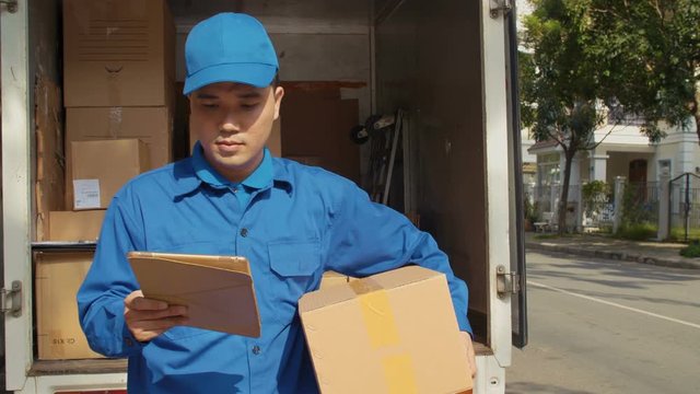 Lockdown of Asian post worker in blue uniform holding tablet in one of his hands, taking parcel from van, then looking at camera and smiling
