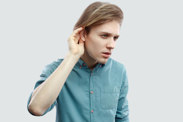 Portrait of serious handsome long haired blonde young man in blue casual shirt standing, holding hand on ear and trying to hear or spy. indoor studio shot, isolated on light grey background.