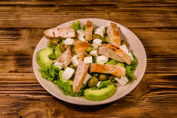 Fresh salad with chicken meat, feta cheese, avocado, green olives and lettuce leaves in ceramic plate on wooden table