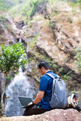 Young man sit and turn back working with laptop at waterfall.
