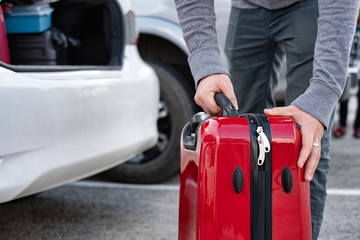 Young traveler loading and picking up red suitcase into truck of car.
