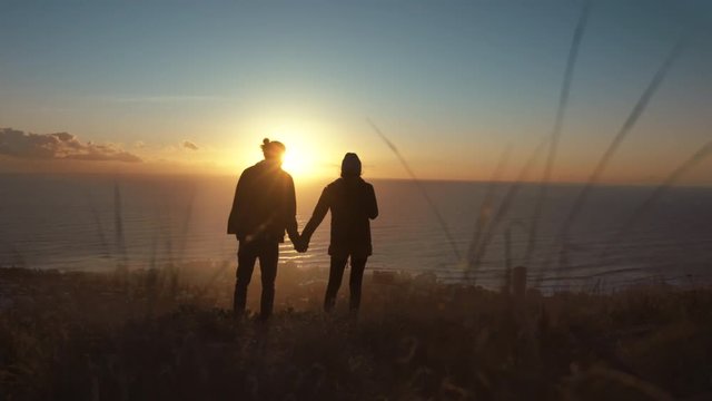 Silhouette of man and woman holding hands and standing on the mountain peak at sunset. Traveling couple on cliff at sunset.