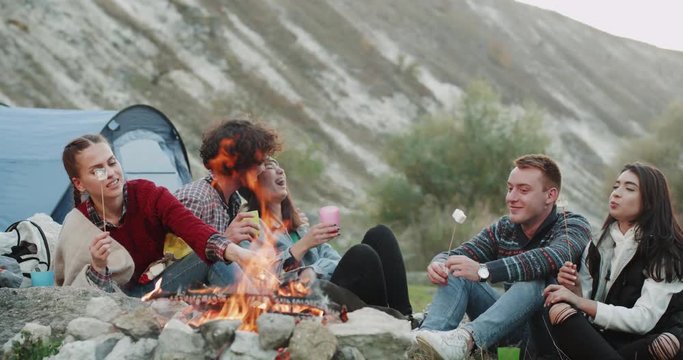 Funny charismatic friends have a great time together at camping , sitting all together at campfire eating marshmallows and drinking wine. 4k