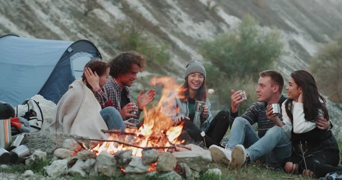 Smiling charismatic friends different races at camping , sitting beside a bonfire and drinking some hot drinks from metallic cups and spending a amazing time together.
