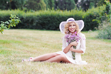 Fototapeta premium Curly blonde in a white hat walks in a park with a bouquet of field violet flowers..