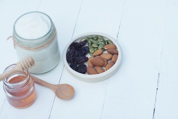 Plain yoghurt in white bowl, honey, cereal, raisin and wood spoon on white wooden table background
