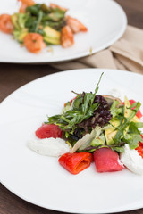 A salad with red fish and greens on a white plate is on the table against the background of another salad.