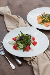 A salad with red fish and greens on a white plate is on the table against the background of another salad.