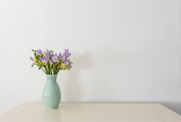 Purple freesia flowers in green vase on stone bench against white wall with copy space (selective focus)