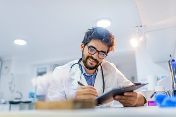 Male doctor sitting with clipboard near window in hospital and filling up medical history form.