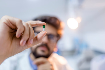 A male doctor in white coat in front of a office, with a stethoscope on one shoulder holding a pill between his fingers