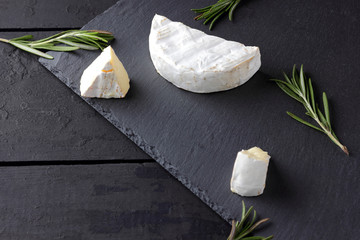 Camembert and rosemary on black stone board. Soft cheese with white mold on black background. Sliced cheese and rosemary branches on slate board. Copy space