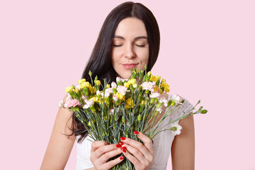Beautiful young lady with straight long hair, smells flowers with pleasant odour, closes eyes, wears white dress, has red manicure, isolated over rosy background. Woman and spring time concept
