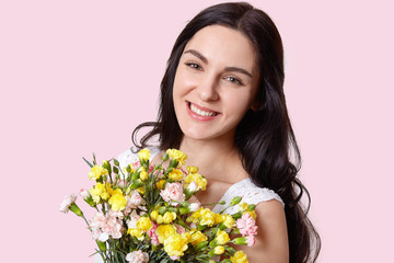 Naklejka premium Portrait of pleasant looking woman with dark hair, has minimal make up, broad smile, recieves flowers on special occasion, isolated over light pink background. Feminity, flowers and beauty concept