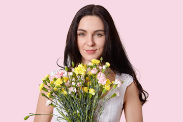 Obraz na płótnie Canvas Close up shot of lovely brunette young woman with healthy soft skin, minimal make up, holds flowers in front, looks at camera with happiness, poses over rosy background indoor. Women and flowers
