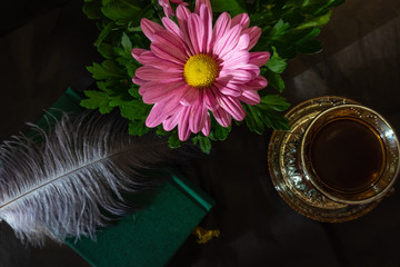 Still life consisting of a cup of coffee, a pink flower, a book and other accessories on a dark background with contrasting light