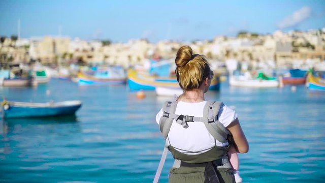 A young mother with a little baby in a sling standing in front of traditional eyed colourful boats Luzzu