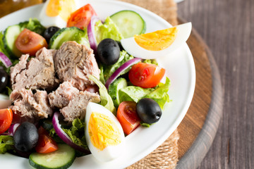 Fresh fish tuna salad made of tomato, ruccola, tuna, eggs, arugula, crackers and spices. Caesar salad in a white bowl on wooden background