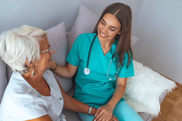 Professional caretaker with her senior charge sitting on a sofa in a bright living room of an assisted living house.