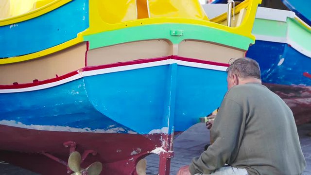 An old man paints traditional eyed colourful boat Luzzu in fishing village Marsaxlokk, Malta