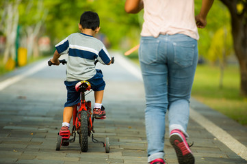 back view lifestyle portrait of mother and young happy son at city park having fun together the kid learning bike riding and the woman running after the child