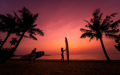 Silhouette of surfers couple holding long surf boards at sunset on tropical beach