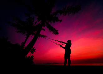 The athlete performs exercises on the beach with fitness straps which are fixed on the palm tree. Sunset