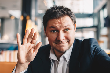 Close-up of good looking young man making a video call in a cafe bar during a break from work, smiling and waving at the camera. Concept of having a long distant relationship.