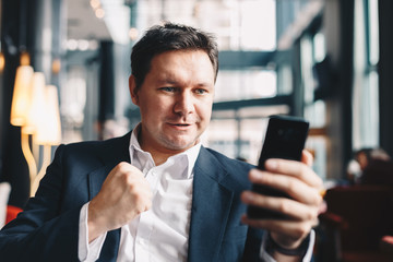 Young businessman sitting in a bar, exulting and raising his hand to celebrate the good financial news he received over his smartphone.