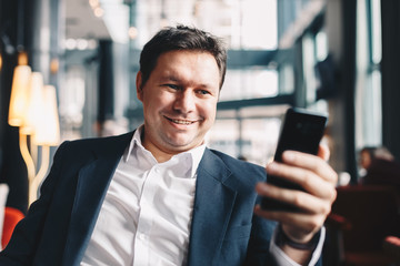 Close up of handsome young man sitting in a bar, smiling while using his mobile phone to text with friends on social media during a break from work.