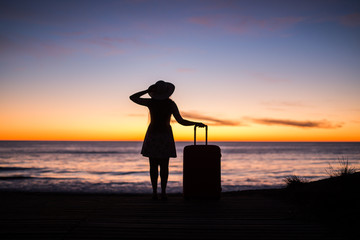Travel, vacation and holiday concept - Young woman standing with suitcase on sandy beach at sea at the sunset