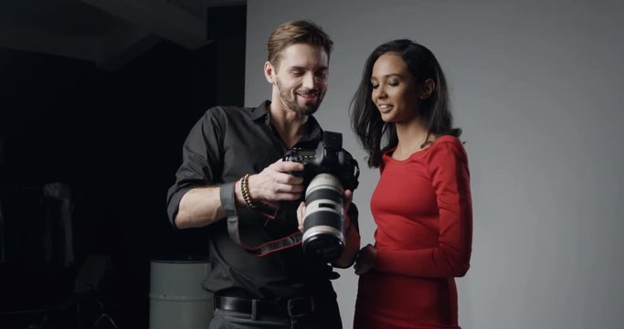 Caucasian man with a photocamera in hands demonstrating pictures just made in the photostudio to the young pretty woman model.