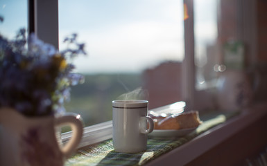 A breakfast consisting of a cup of hot coffee and a plate with a glazed cake on the elbow-board of a terrace lit by the bright rising sun, with a vase of flowers in the foreground.