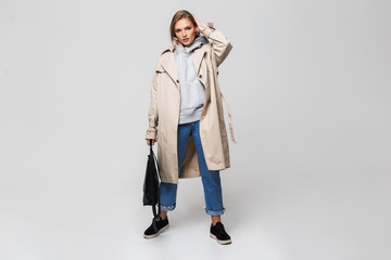 Young beautiful woman in trench coat and jeans holding black backpack in hand while dreamily...