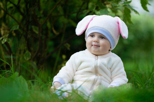 Child girl with chubby cheeks in bunny suit seating on grass in forest.