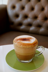 Cup of fluffy milk foam hot cappuccino coffee on white table with blurry sofa in background 