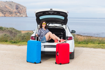 Young woman sitting in back of car smiling and getting ready to go. Girl sitting in the open trunk of a car. Summer road trip