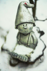 Winter Goblin. A small garden gnome or dwarf. He sits and looks up into the camera. Snow covers him but he acts to peace. A cute little guy with a pointed hat