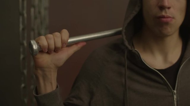 Aggressive grim looking street hooligan in hoodie holding baseball bat on shoulder, expressing hostility and danger in foggy interior. Violent criminal guy with baseball bat ready to attack victim.