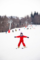Little smiling girl in red overall training skiing