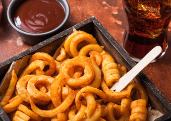 Curly fries fast food snack in wooden box with ketchup and glass of cola on rusty stone kitchen background. Unhealthy junk food