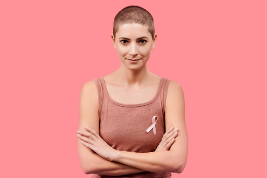 Smiling mid 30s woman, a cancer survivor, wearing pink breast cancer awareness ribbon, isolated over living coral background. Support, solidarity, screening and prevention concept.