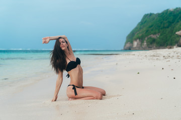 Sexy brunette girl in black swimwear sits and sunbathes on ocean beach