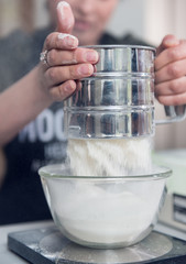 A woman sieving white baking flower making it into a fine refined  batch, preparing for baking cake recipe. Metal flour serving.