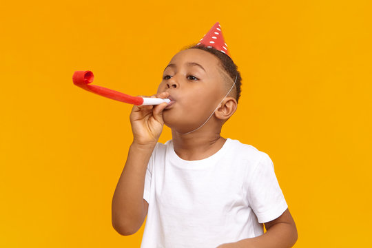 Happy Afro American little boy celebrating his ten year birthday with friends, posing isolated at blank yellow wall, blowing whistle, holiday hat on his head. Fun, joy, celebration and holiday