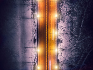 Aerial top down view of illuminated bridge road with motion blurred traffic car lights on background of frozen river in ice with snow. Abstract winter drone photo