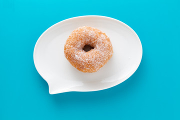 A doughnut coated with sugar on plate shaped like text bubble, on blue background.